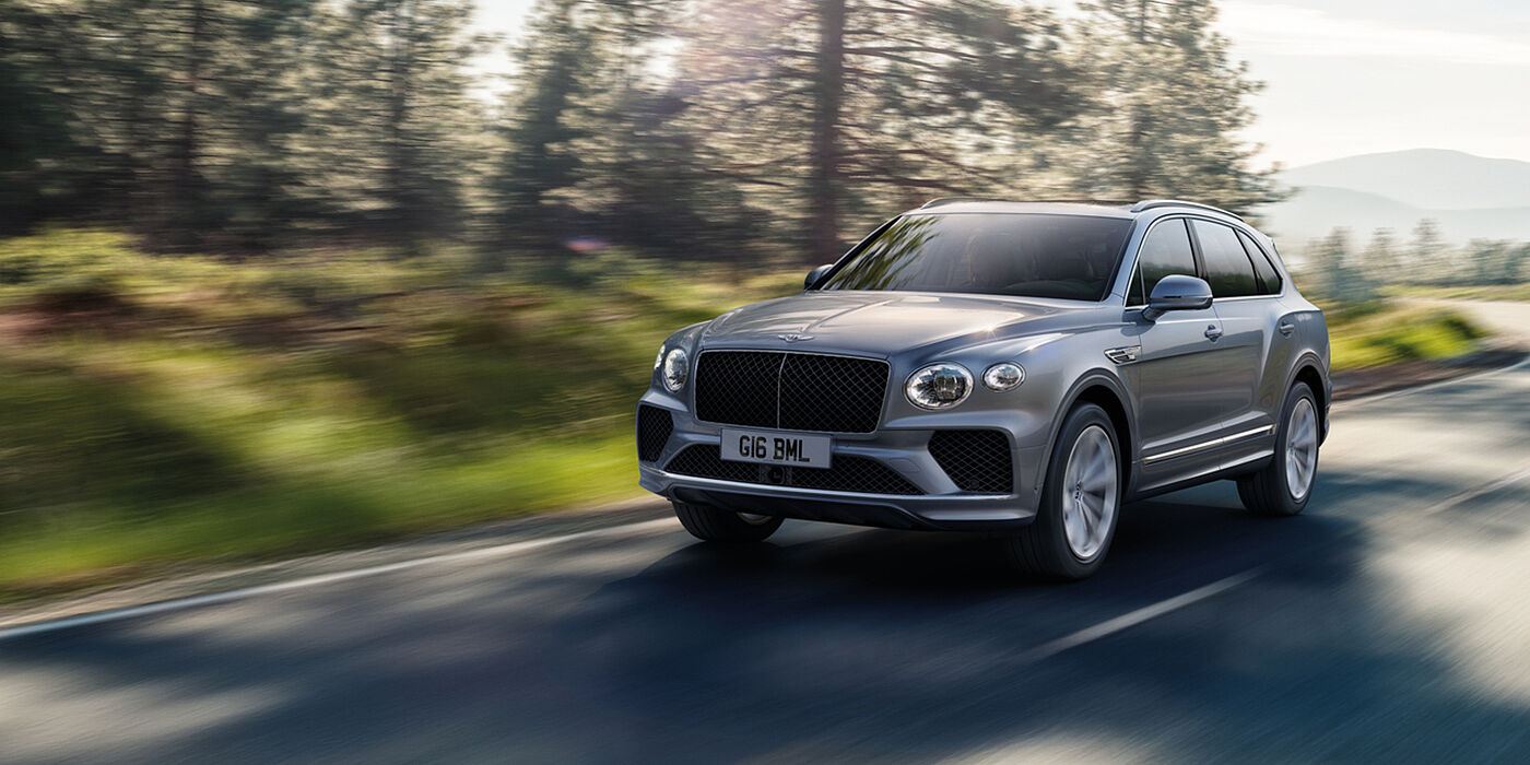Bentley Manila Bentley Bentayga luxury SUV front three quarter in Moonbeam paint, driving on an country road past trees.