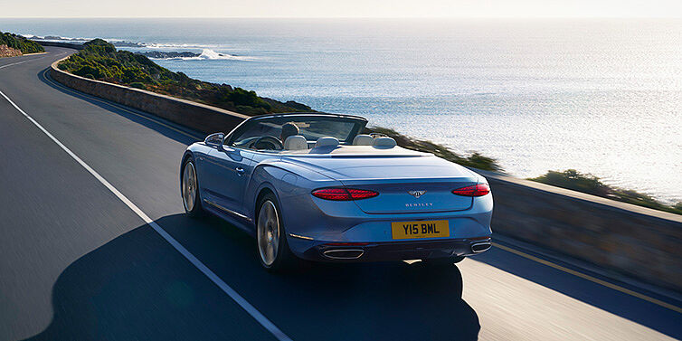 Bentley Manila New Bentley Continental GTC Azure convertible rear view in Light Windsor blue paint with 22 inch ten spoke wheels driving dynamically on an ocean road in the sunshine