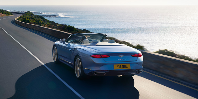 Bentley Manila New Bentley Continental GTC Azure convertible rear view in Light Windsor blue paint with 22 inch ten spoke wheels driving dynamically on an ocean road in the sunshine